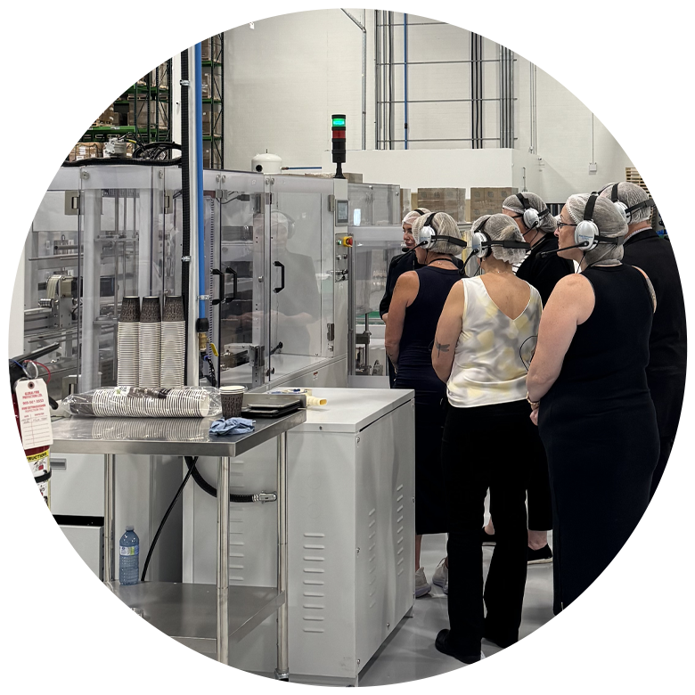 Circular photo of visitors wearing hairnets and audio headsets on a guided tour of EcoGuardian's Aurora facility, standing beside a cup-manufacturing machine with stacked paper cups on a stainless steel table.